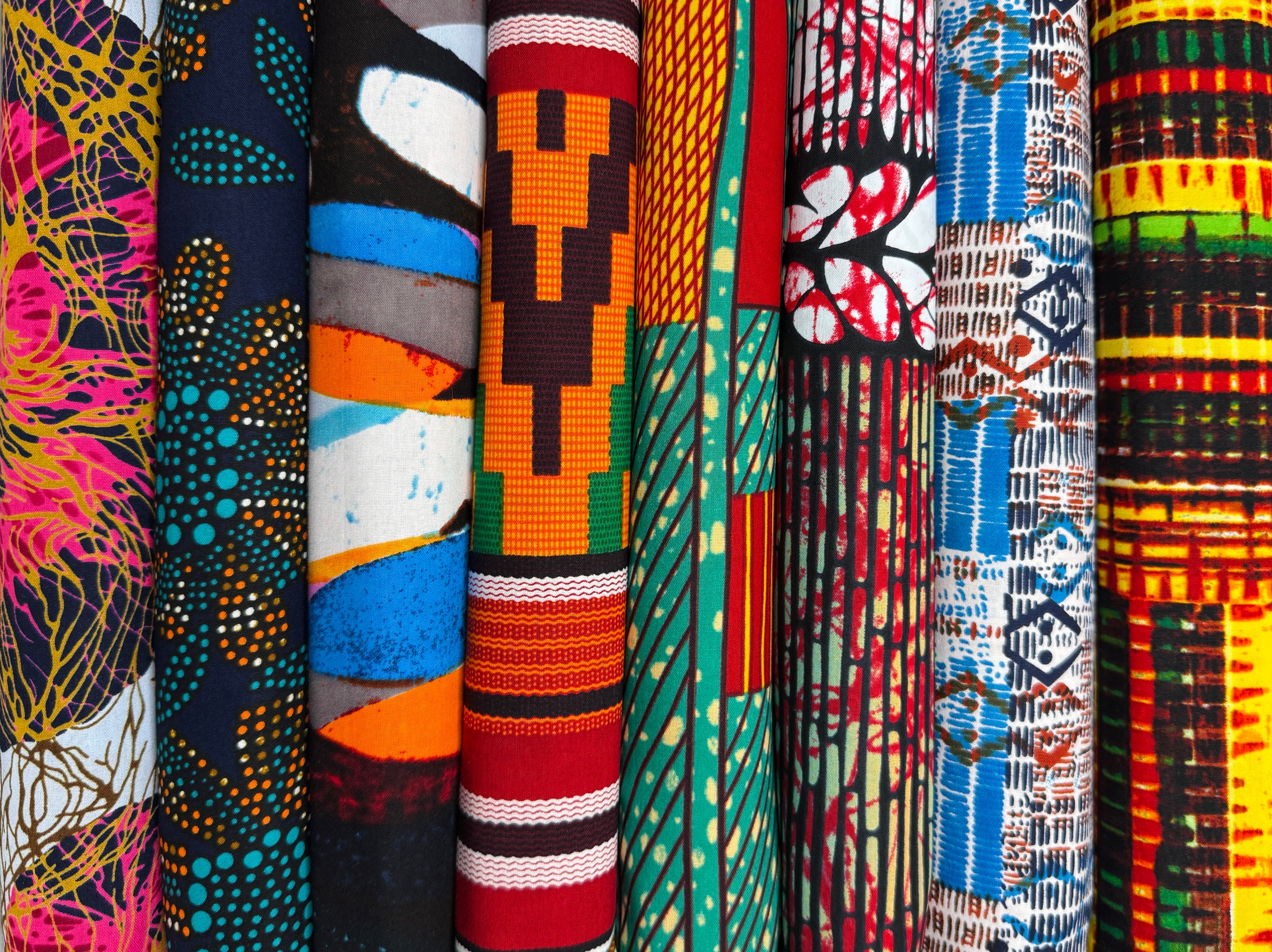 A vibrant spread of African fabrics by the yard, featuring bold Ankara wax prints, Kente motifs, and tribal patterns in a spectrum of colors, celebrating West African textile heritage.